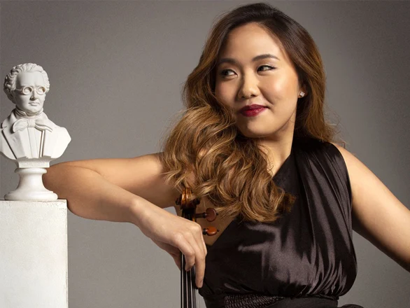 Stella Chen sits and looks over her shoulder while holding the top of a violin. Her arm rests in front of a classical statute bust.