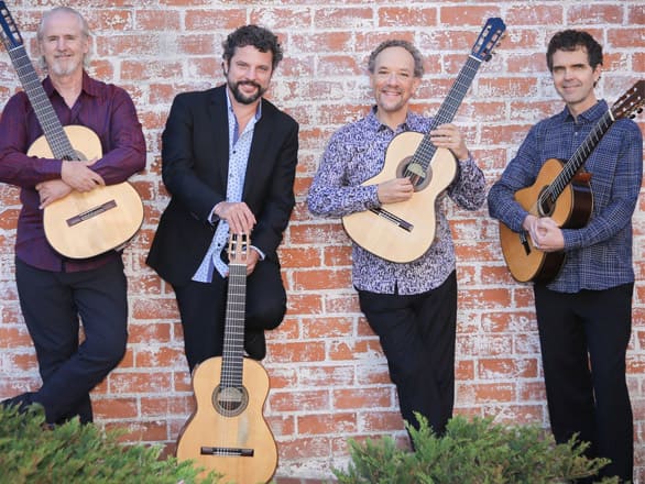 The Los Angeles Guitar Quartet hold their guitars and pose for a publicity photo in front of a brick wall.
