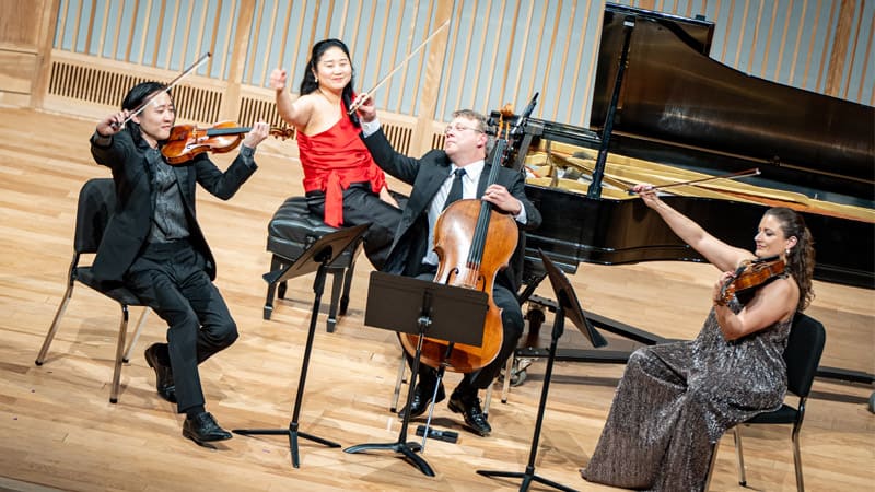 Four classical musicians perform on stage — two violinists, a cellist, and a pianist — enthusiastically raising their bows in a celebratory gesture. The pianist in a red dress smiles while seated at the grand piano, and the string players, dressed in formal attire, are positioned around music stands on a wooden stage.