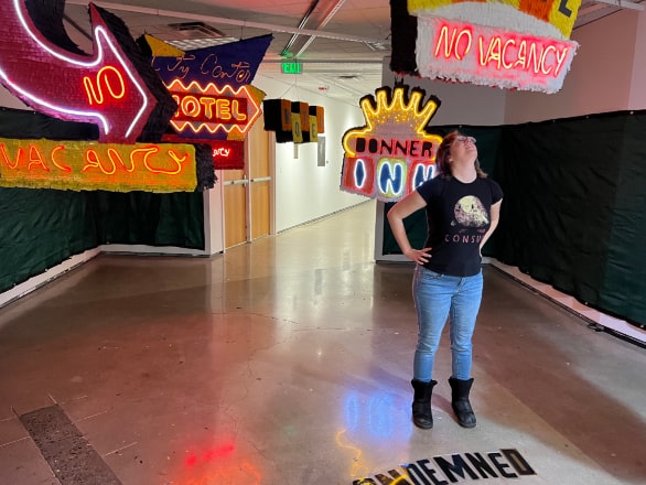 anna newman standing in hallway with neon signs saying "no vacancy" 