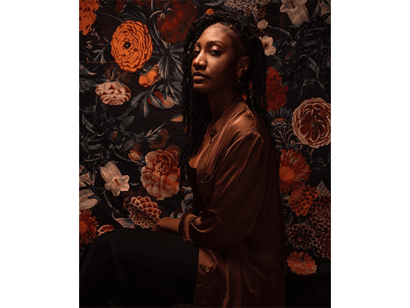 Image of a dark-skinned person wearing a brown jacket against floral wallpaper