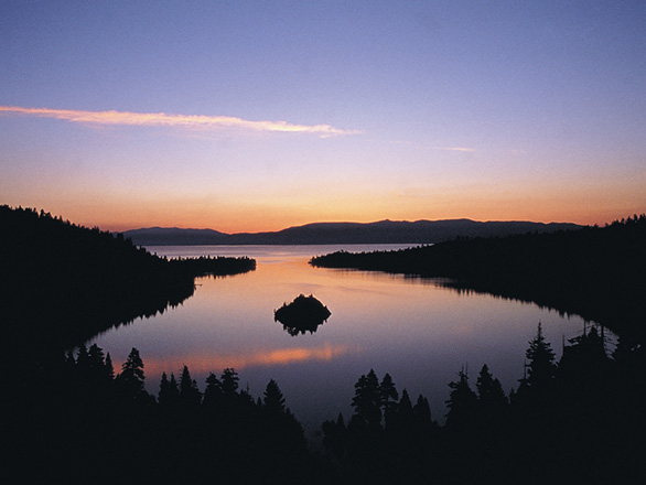 A sunset falls over the calm waters of Lake Tahoe, with trees surrounding the lake with purple and red hues filling the sky.