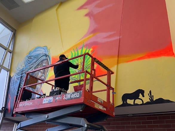 Artist Edwin Martinez-Escobar stands on a red SkyJack lift while painting a vibrant indoor mural. The artwork features a stylized blue-haired face, a green and yellow abstract burst, and a horse silhouette with desert plants, all set against a gradient yellow-to-red background.