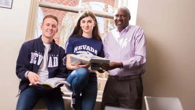 Two students wearing Nevada branded jackets and shirts hold books and stand next to a professor who is also holding a book.