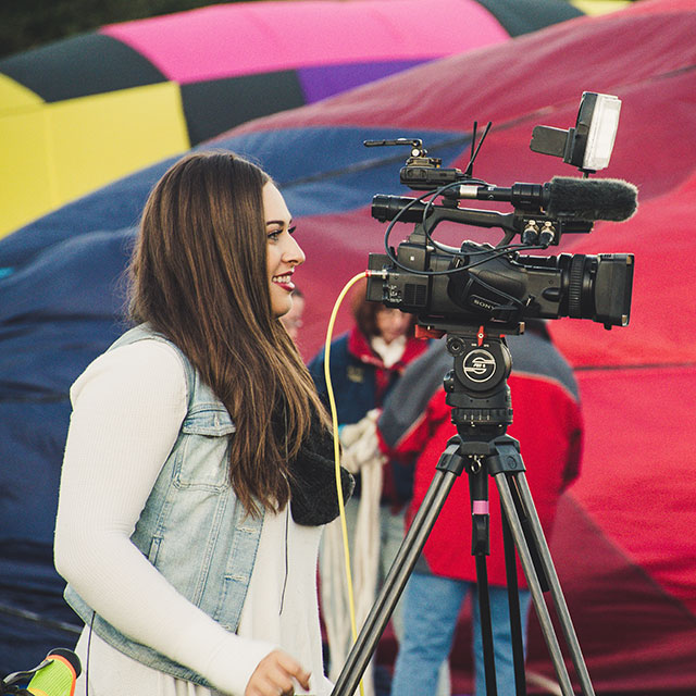 Student stands behind video camera interviewing news subject
