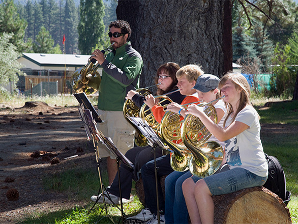 Students play instruments outside while sitting on a log.