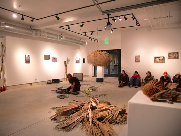 An art gallery, with textiles on the floor and an artist in the middle of the space. Spectators sit along the edges.