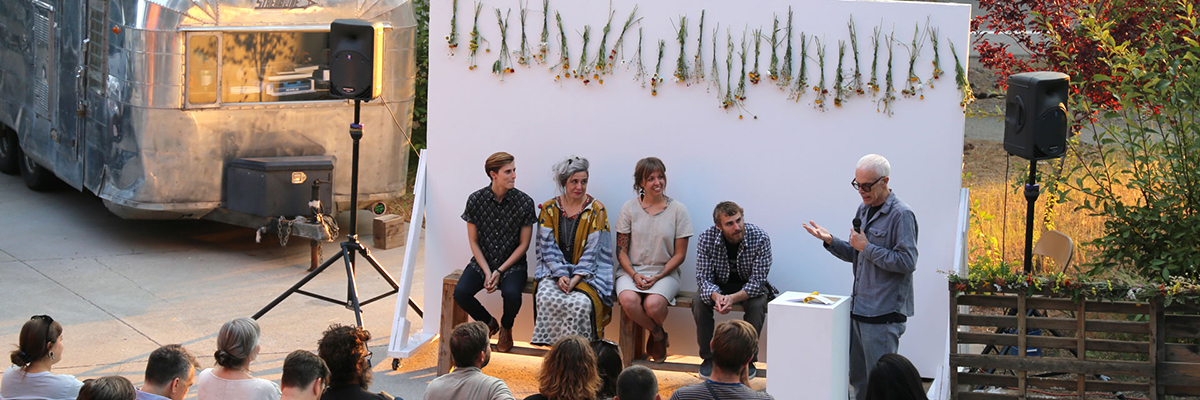 An outdoor panel discussion with a flower-decorated backdrop and airstream trailer.