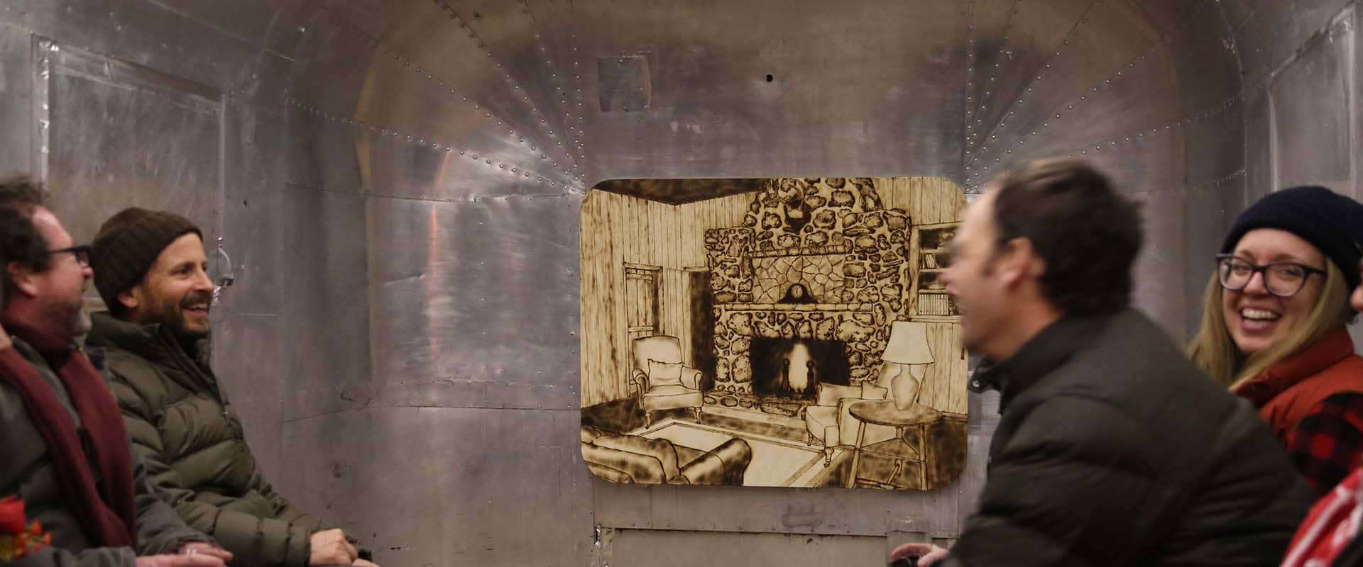 People wearing winter jackets sit smiling and talking in a space with wood-burned artwork and metal walls.