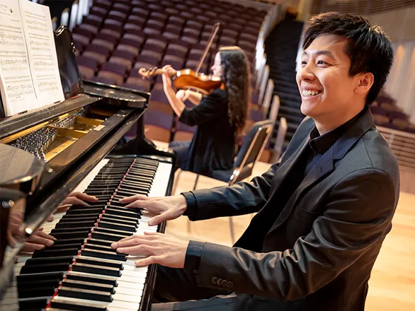 A person wearing a suit is seated and playing a piano while a person seated behind them plays a violin in a large, multi-seat and empty theatre.