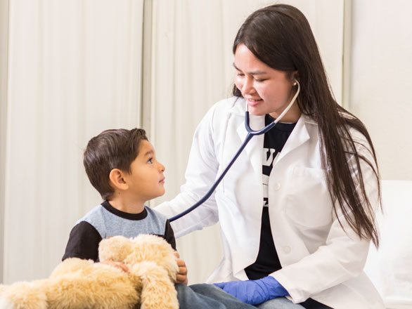 A student wearing a white coat sits on a hospital bed with a child and listens to his heart. There is a stuffed animal in the foreground.