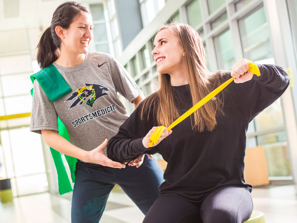 A student wearing a Wolf Pack sports medicine shirt guides another student using a resistance band to exercise her arms