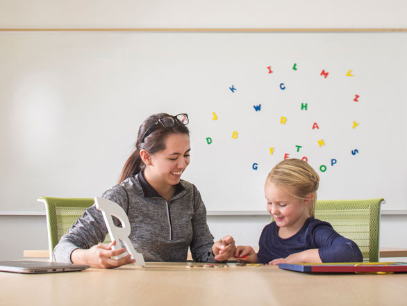 A student in a gray shirt holding up a letter R sitting next to a child, and they are both placing colorful letters on the table.