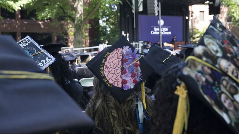 Graduates seated at the University of Nevada, Reno Spring 2026 Commencement ceremony wear decorated graduation caps while facing the stage outdoors. One cap in the center features a painted brain design surrounded by flowers and the phrase “You’re Gonna Go Far.”