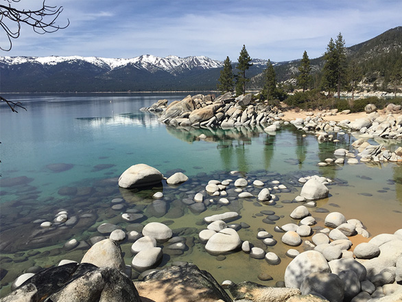 A rocky beach lines Sand Harbor at Lake Tahoe, a giant blue lake with trees lining the lake and mountains in the distance.