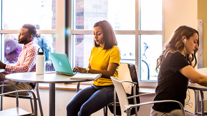 A student wearing a yellow shirt works on a laptop at a table, flanked by two other individuals working on laptops on tables on either side.