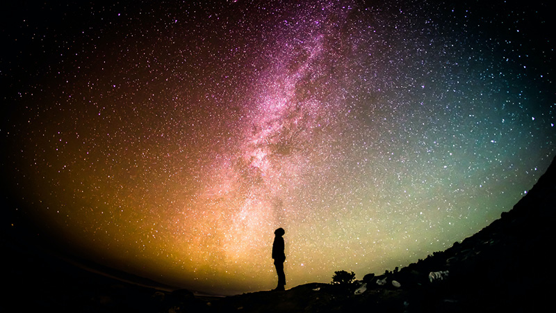 Person staring into the night sky at a sea of swirling colors, from pink to orange, to blue and green.