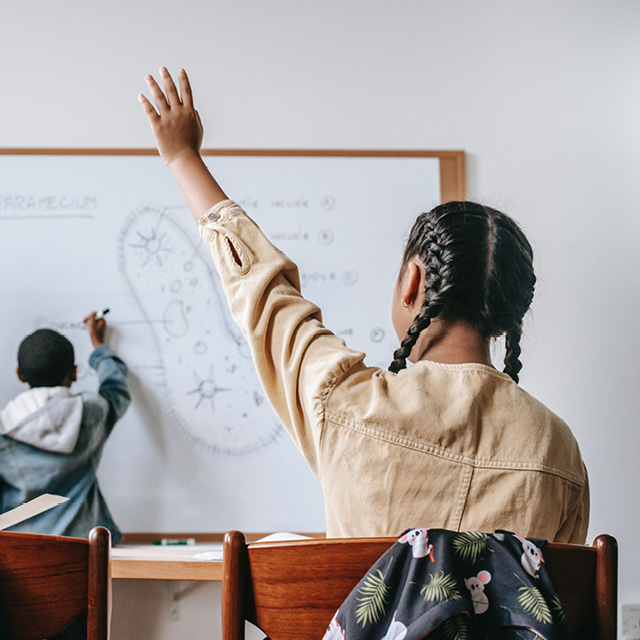 Female student raising hand in classroom, as another student writes on a whiteboard. 