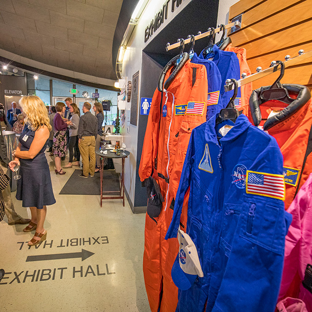 Visitors near the entrance of the Exhibit Hall, congregating and talking amongst themselves, as space suits hang on a rack to the right.
