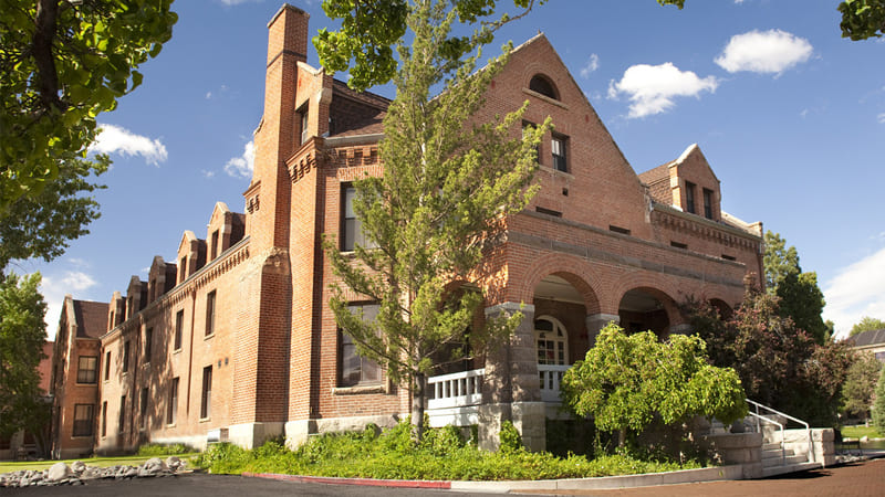 An image of Manzanita Hall, a brick covered building with steps leading to double doors and trees and bushes lining the building