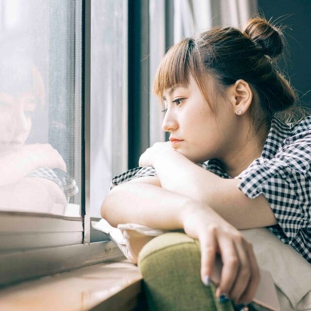 Young woman of Asian decent looking out the window sadly