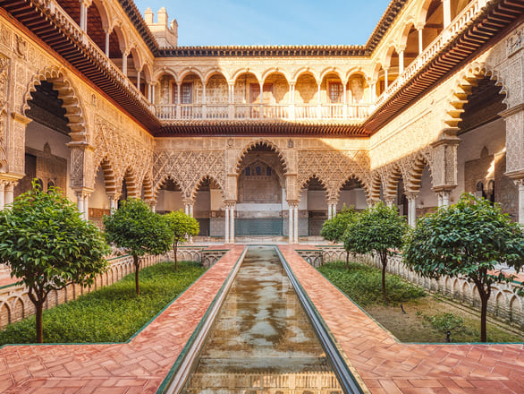 Ornate Moorish-style courtyard with a central reflecting pool, surrounded by detailed arches and columns. Lush greenery adds vibrant contrast.