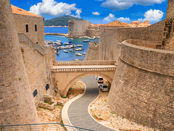 A view of ancient stone walls with an arched bridge, overlooking a tranquil harbor filled with boats and lush green hills in the background.