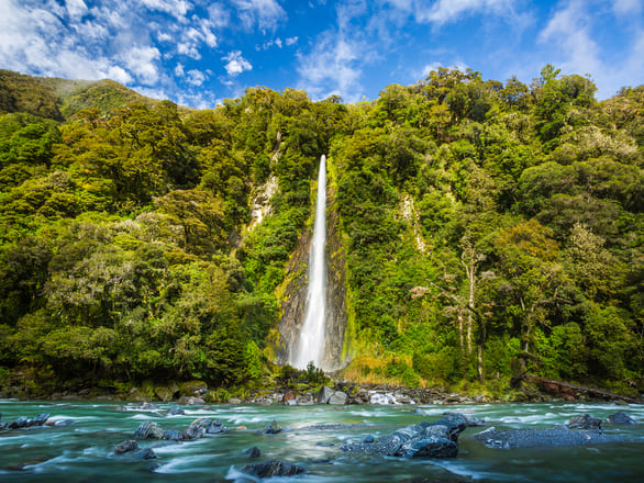 A serene waterfall cascades down a lush green hillside into a flowing river, under a bright blue sky. The scene conveys tranquility and natural beauty.