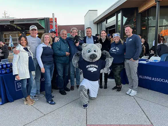 A group of Nevada fans stand with University President Brian Sandoval during a Pack Midtown event. Wolfie Jr. mascot kneels in front of the group of people posing for the photo.