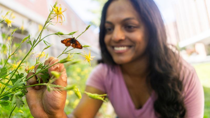 Woman smiling, looking at flower with monarch butterfly on it.