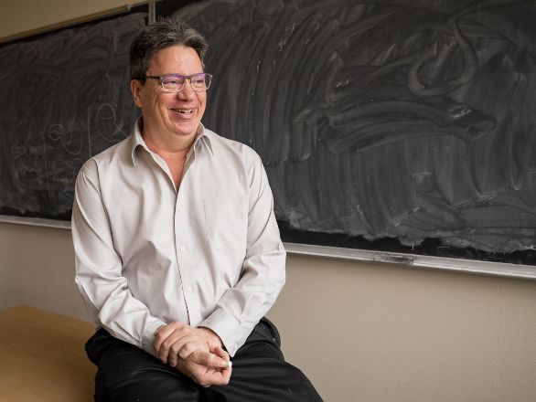 Professor sitting in front of chalkboard smiling.