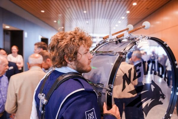 A band member in uniform plays a large drum labeled "Nevada" during an indoor event with attendees in the background.