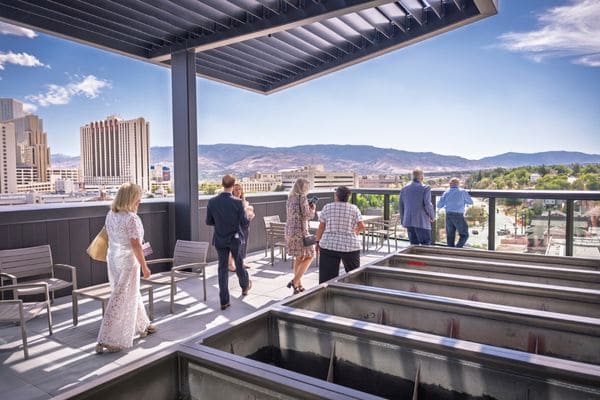 People stand on a rooftop terrace under clear skies, some chatting and others enjoying the city view.