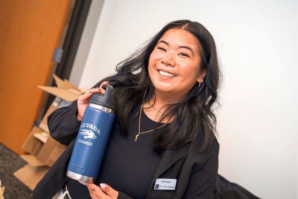 A person smiles while holding a blue "Nevada Business" water bottle indoors, with stacked boxes in the background.