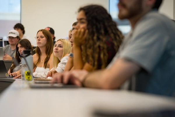 Students sit closely together at desks, attentively focused on something outside the frame during a classroom session.