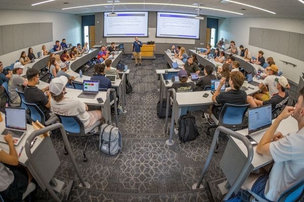 A panoramic view of a classroom where students use laptops at desks while an instructor presents slides on dual screens.