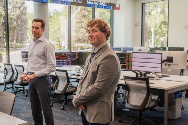 Two individuals in business attire stand in a financial lab filled with monitors displaying stock data.