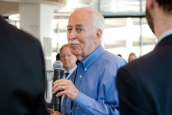 An older man speaks into a microphone at an indoor event, with others nearby listening attentively.