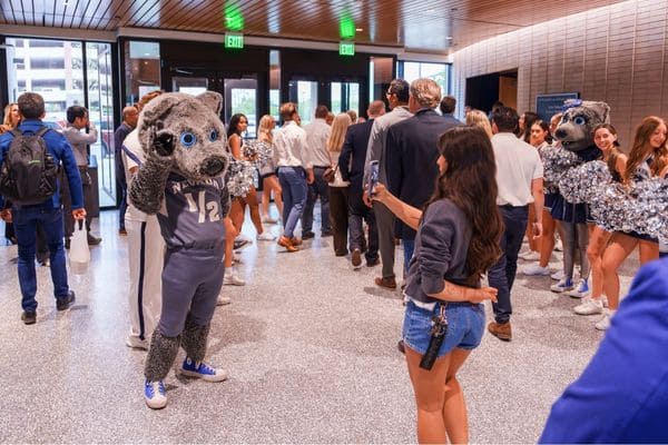 A wolf mascot interacts with attendees at an indoor event, surrounded by cheerleaders and other participants.