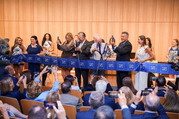 A group of people participate in an indoor ribbon-cutting ceremony, holding scissors to cut a blue ribbon with "N" logos as an audience watches.