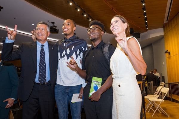 Four people pose together indoors, three making hand gestures and all smiling at the camera.