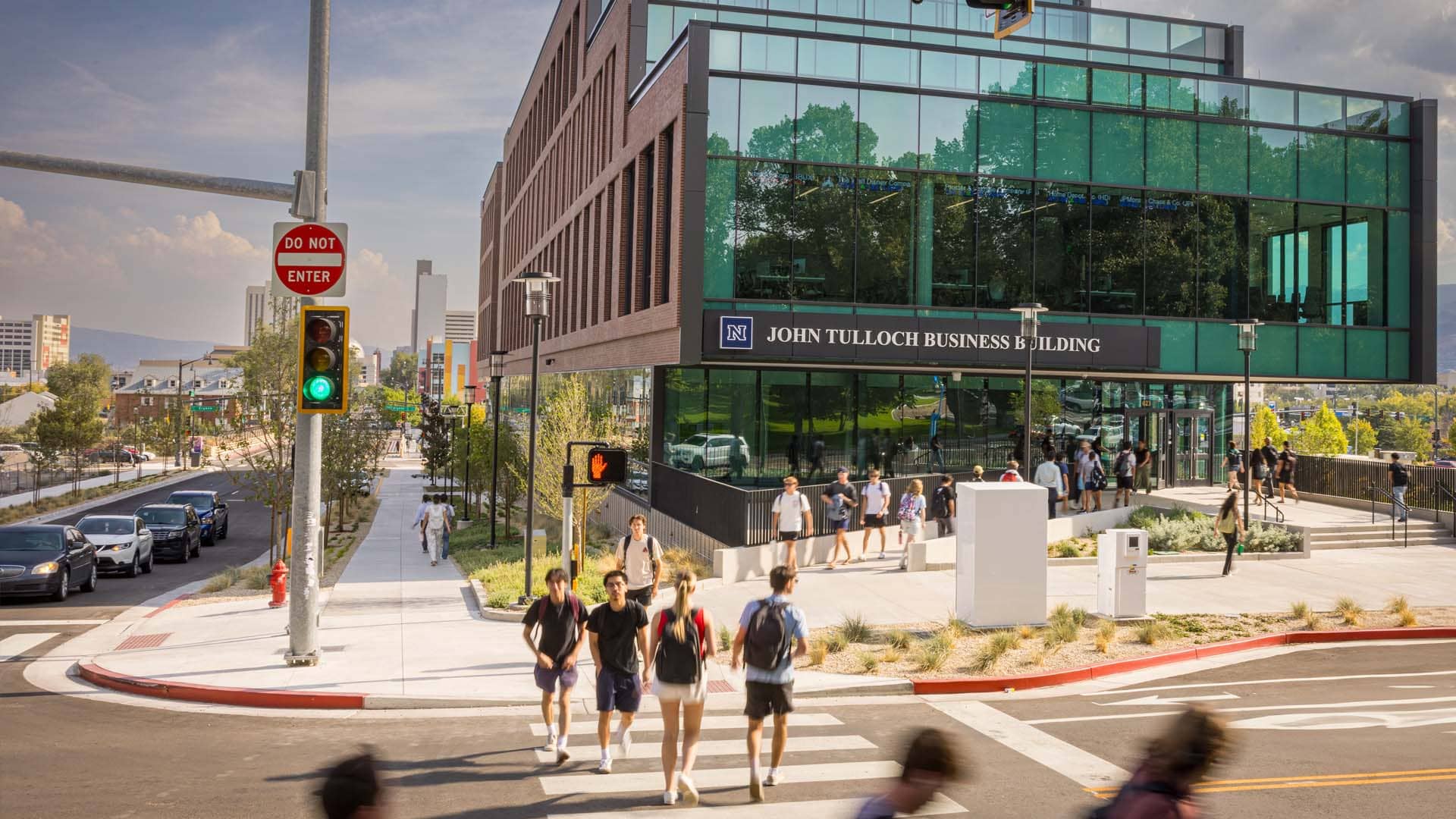 A modern multi-story building labeled "John Tulloch Business Building" stands at a busy intersection with a crosswalk where pedestrians are walking. The structure features large glass windows and contemporary architecture. A traffic light shows green, and a "Do Not Enter" sign is visible on a nearby pole. Surrounding the building are trees, other campus structures, and distant mountains under a partly cloudy sky.