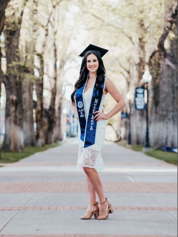 A woman in graduation attire stands on a tree-lined path, wearing a blue stole reading “NEVADA.”