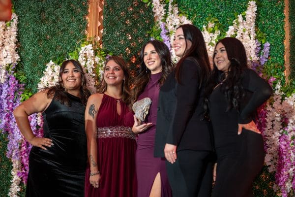 Five women in formal attire posing in front of a floral green backdrop.
