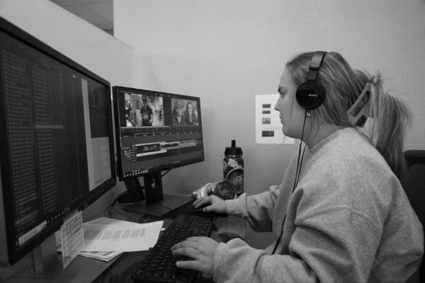 A woman using dual monitors, wearing headphones, at a desk in an office. 