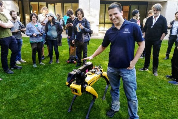 A man smiles and poses with a yellow quadruped robot outdoors, surrounded by a group of people observing and using mobile devices.