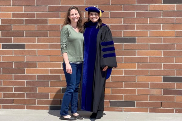 Vanessa Gutierrez, wearing a black doctoral gown with blue velvet panels and a matching tam, stands smiling next to her mentor, Dr. Cyprian Rosetto, who is dressed casually in a green shirt and jeans.