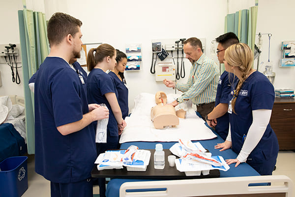 A clinical training session with five individuals in blue medical scrubs and one instructor in a striped shirt gathered around a hospital bed. The instructor demonstrates a procedure on a medical training mannequin. A table beside the bed holds medical supplies such as gloves, syringes, and bottles. The room features medical equipment on the walls and partially drawn privacy curtains, indicating a hands-on learning environment for healthcare professionals or students.