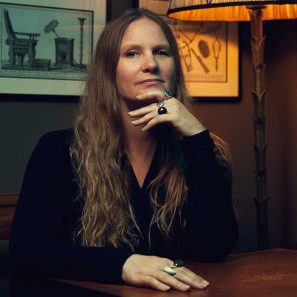 Lidia Yuknavitch long hair seated at a table, appearing engaged in thought or conversation.