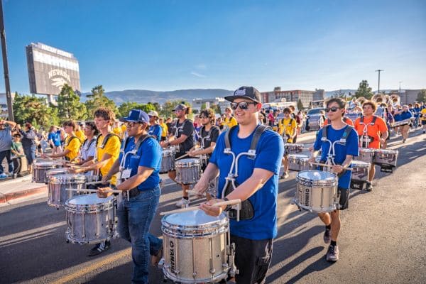 Members of a college marching band dressed in different colored shirts play the drums.
